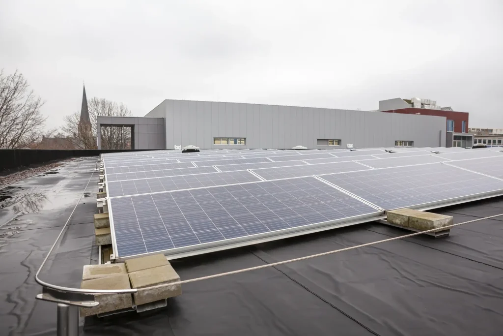 Flachdach-Photovoltaikanlage auf schwarzer Dachabdichtung, die ohne Dachdurchdringung mittels Ballastierung durch schwere Betonsteine auf den Metallschienen gesichert ist.
