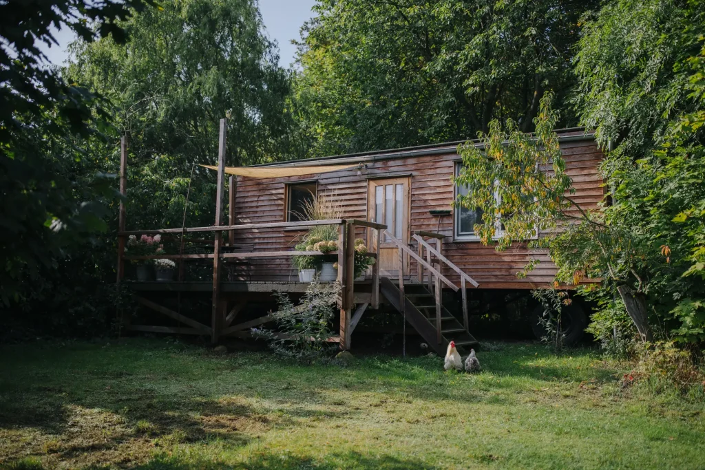 Ein kleines, gemütliches Tiny House aus Holz in einem Garten, umgeben von üppigem Grün und Bäumen. Vor dem Haus befindet sich eine erhöhte Holzterrasse mit Blumenkübeln und einem Sonnensegel. Eine kleine Treppe führt zur Eingangstür. Auf der Rasenfläche vor dem Haus laufen zwei Hühner umher.
