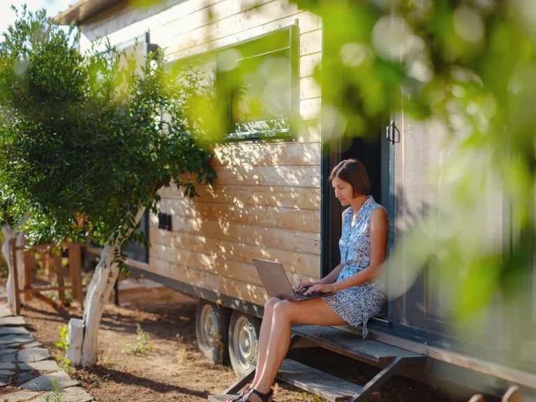 Eine Frau sitzt auf den Stufen vor einem kleinen, mobilen Tiny House aus Holz und arbeitet an einem Laptop. Sie trägt ein sommerliches, blau gemustertes Kleid. Das Tiny House steht auf einem Anhänger mit zwei Achsen und ist von Bäumen und grünem Laub umgeben, das den Vordergrund leicht verschwimmen lässt. Der Boden ist trocken und von vereinzelten Pflanzen bewachsen, während ein gepflasterter Weg links vorbeiführt.